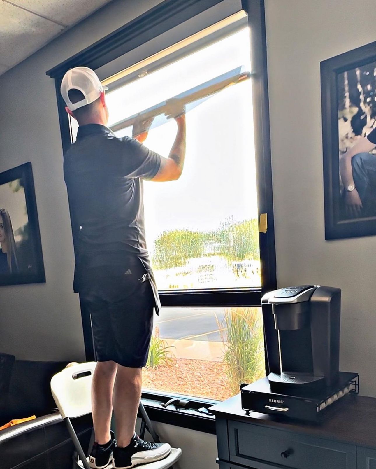 A person standing on a small stepladder installing a window shade in an office or home setting.