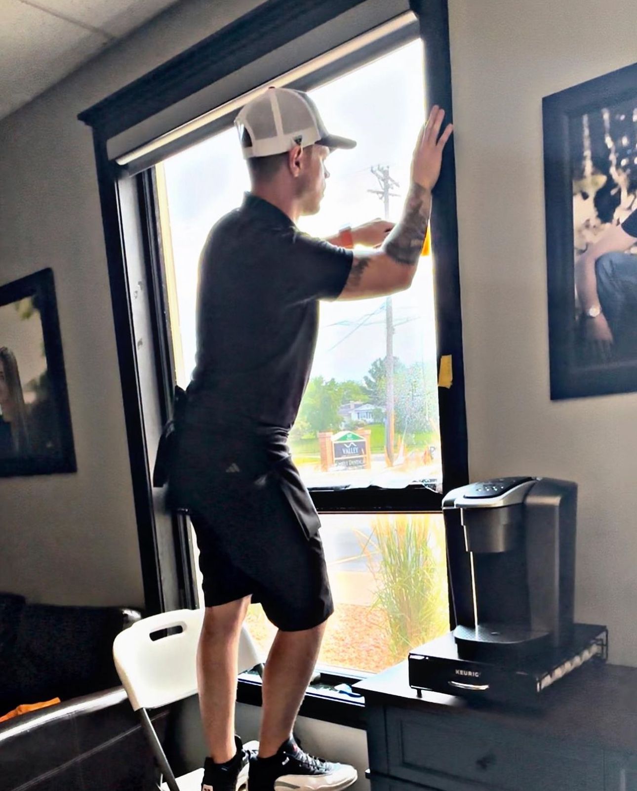 A person in a baseball cap stands on a chair, applying dark film to a window in a room with framed wall art.