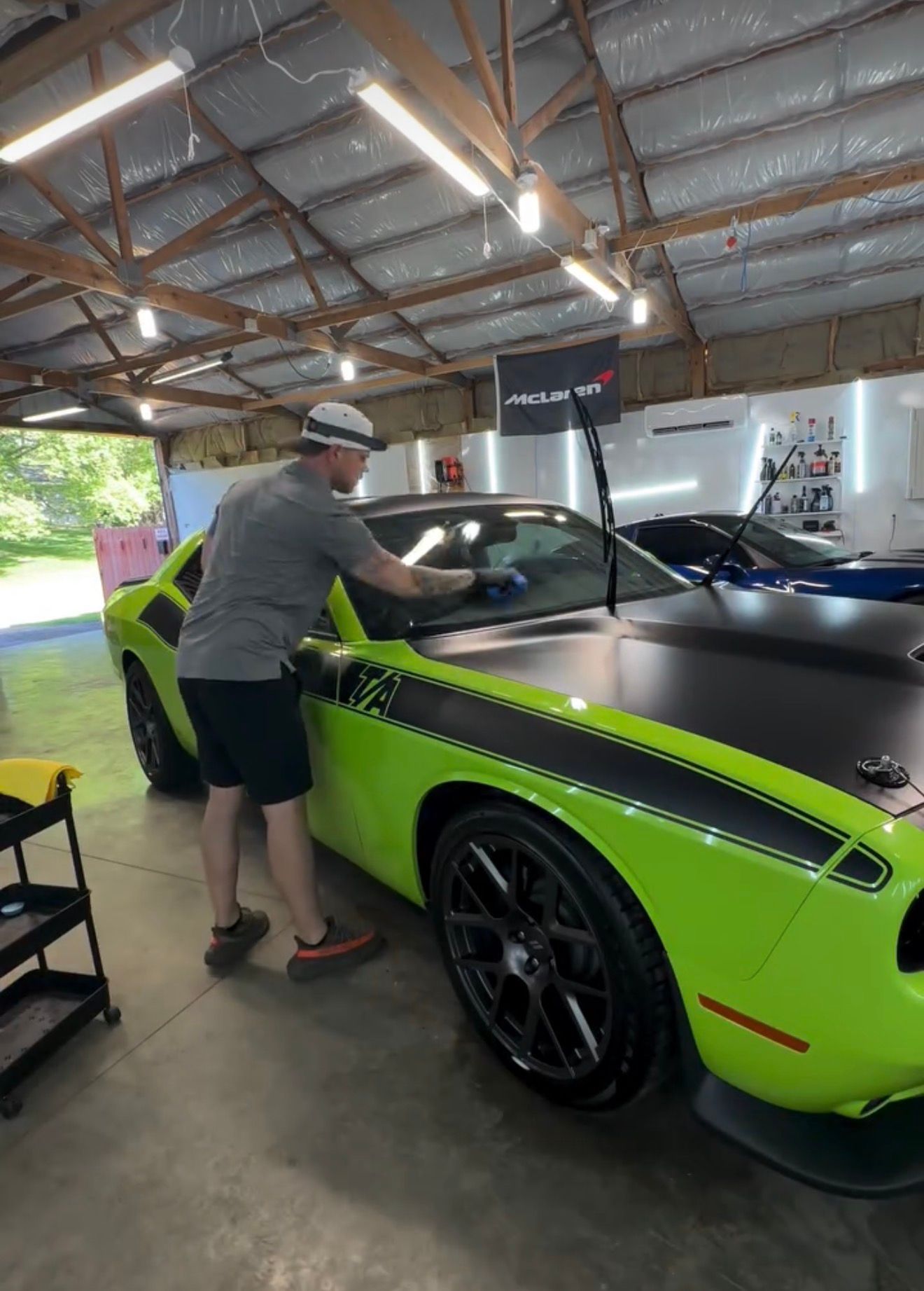A person in a gray shirt and shorts cleans the windshield of a lime-green Dodge Challenger in a well-lit auto shop.