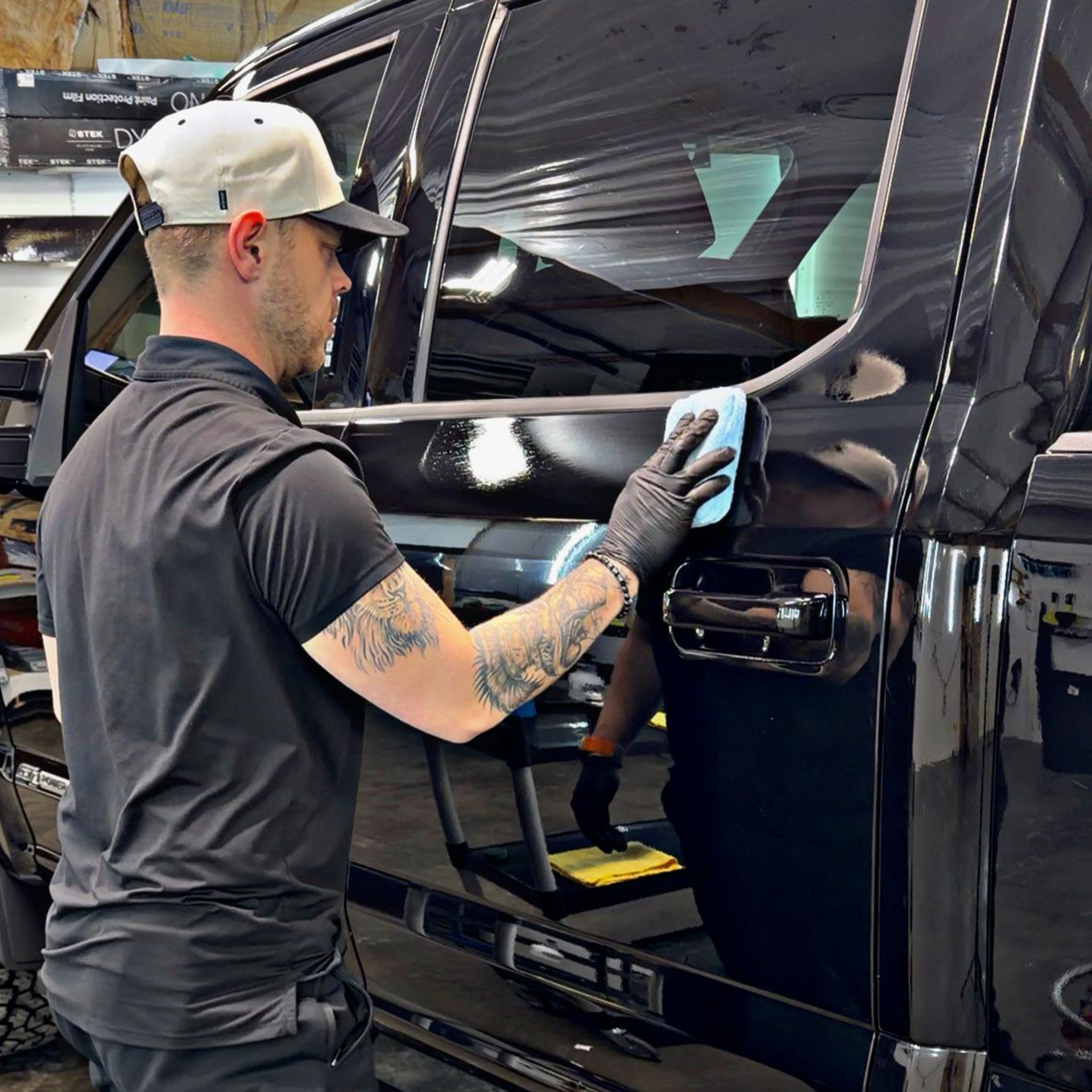 A person wearing a hat and black clothing wipes the side of a black truck with a blue cloth in a workshop.