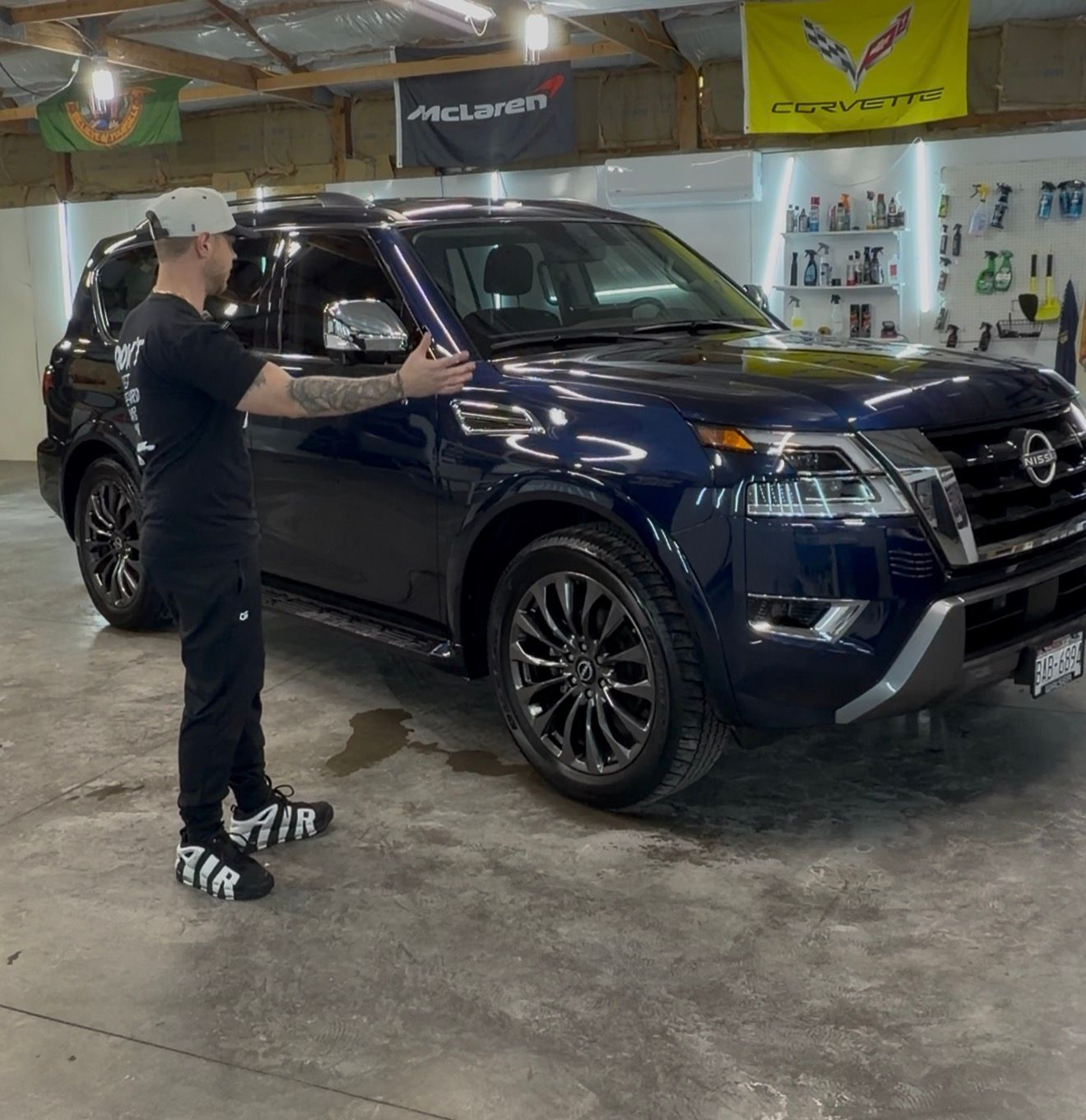 A person gestures toward a dark blue Nissan Armada SUV inside a workshop with automotive gear on the walls.