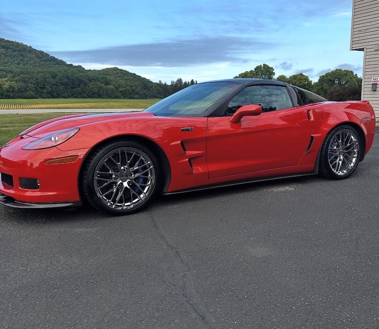 A bright red C6 Chevrolet Corvette Z06 sports car parked on an asphalt driveway with a mountain backdrop.