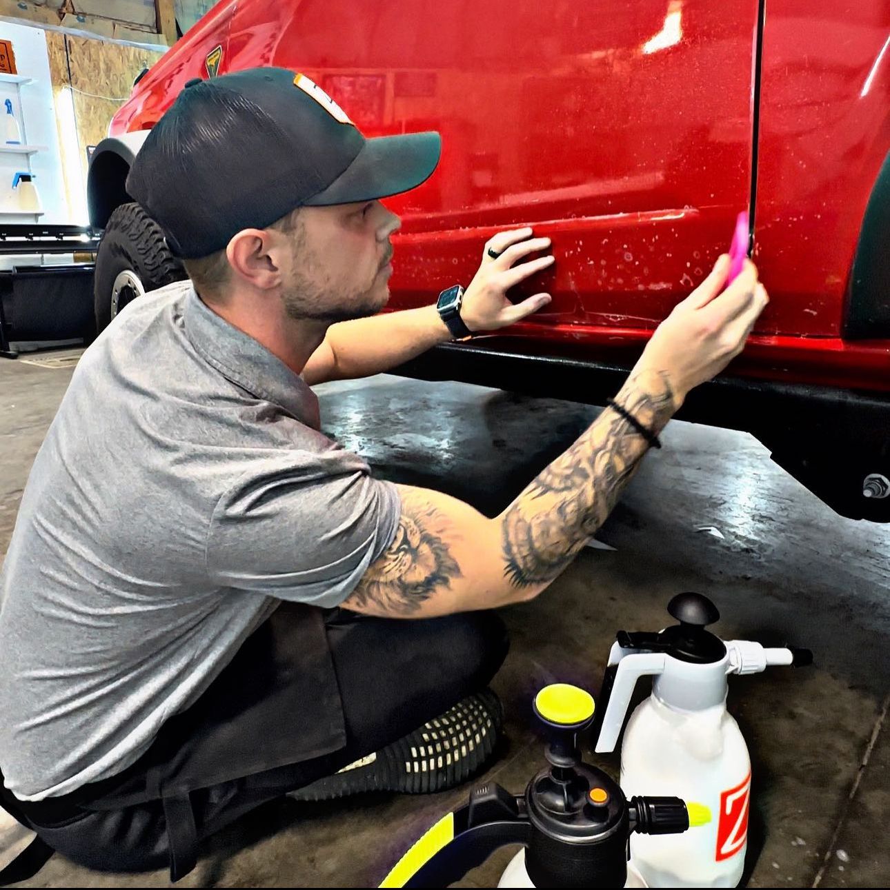 A person in a garage uses a pink squeegee to apply clear protective film to the red side panel of a vehicle.