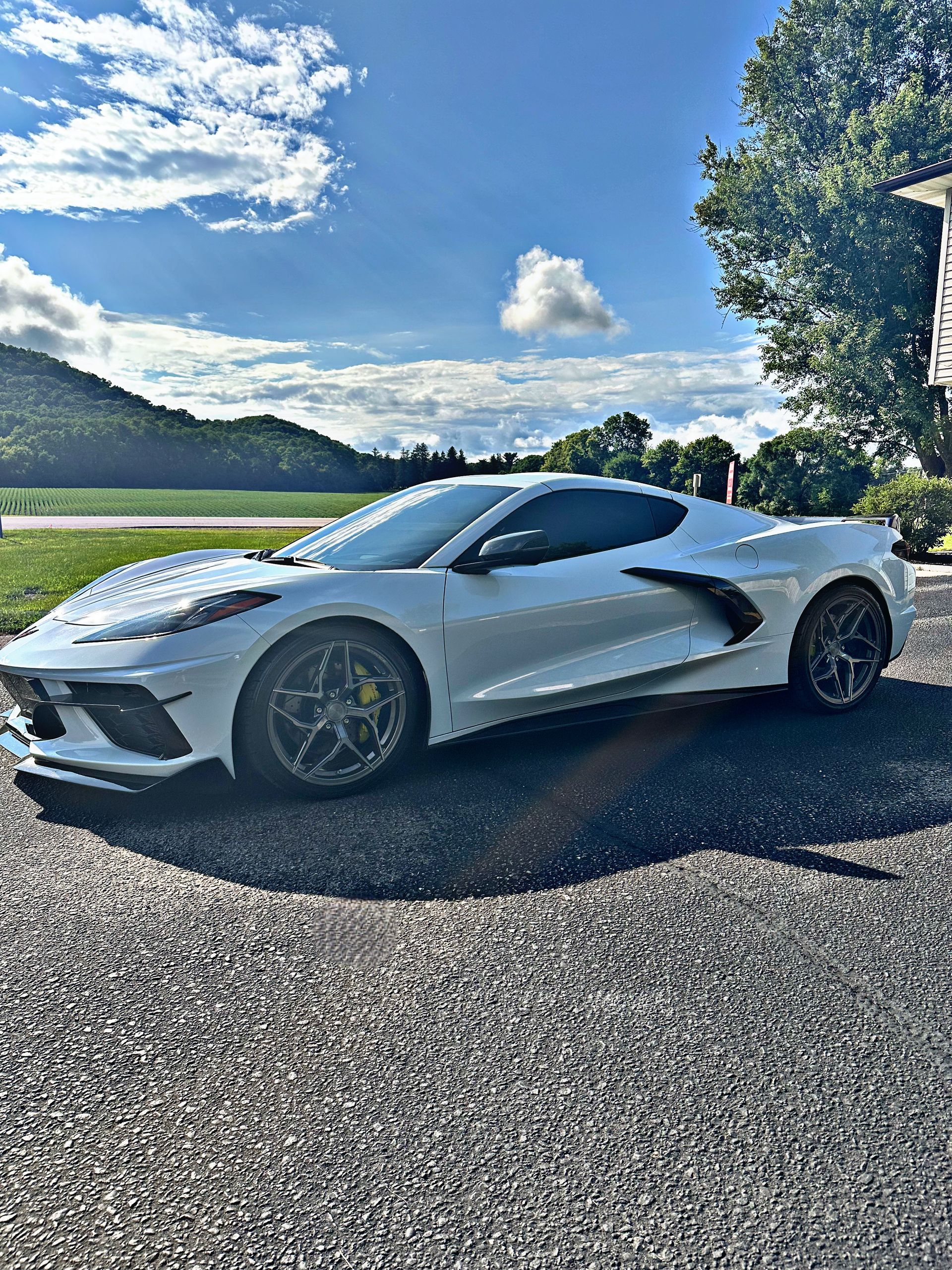 A white Chevrolet Corvette C8 sports car parked on a gravel surface under a bright blue sky with scattered clouds.