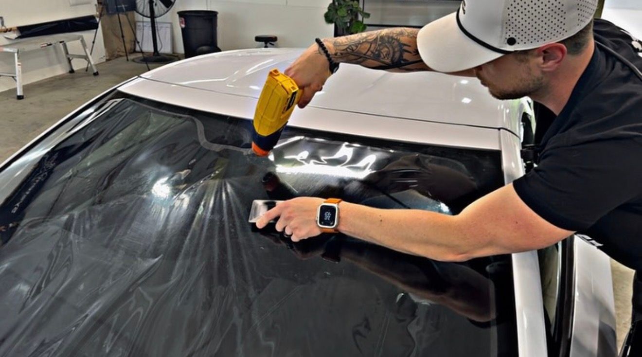 A person uses a yellow heat gun to apply dark window tint film onto the windshield of a white vehicle in a workshop.