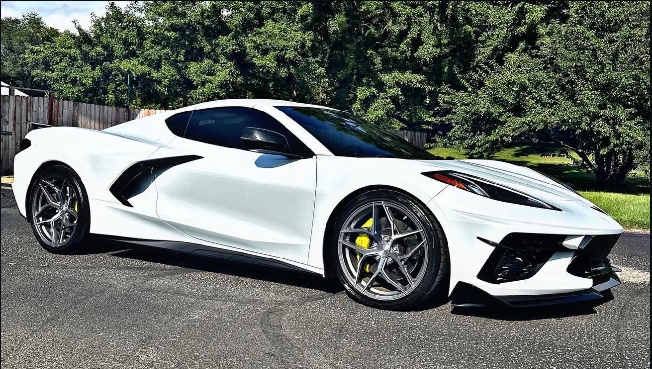 A white Chevrolet Corvette C8 sports car parked on an asphalt driveway with green trees in the background.