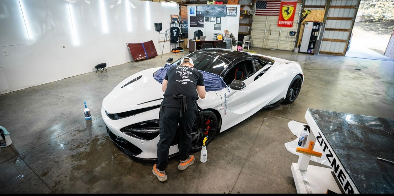 A mechanic works on a white sports car inside a bright, clean automotive shop with a Ferrari logo on the wall.