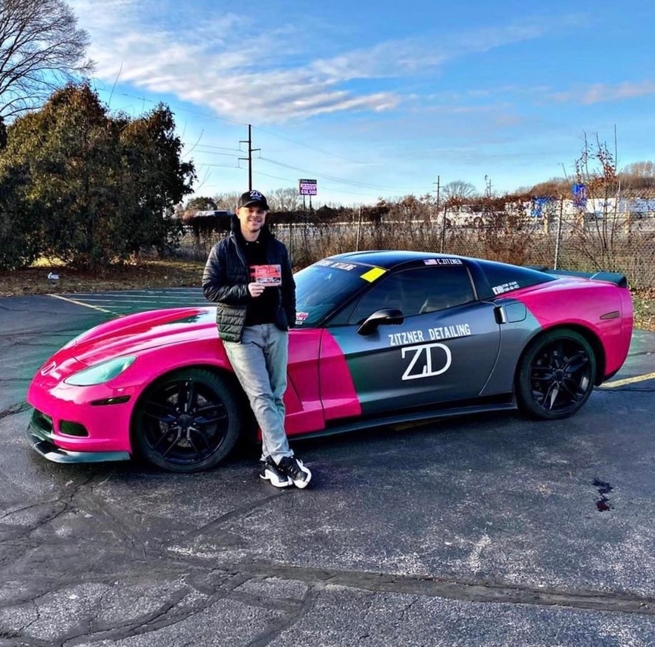 A person wearing a cap and jacket stands beside a two-toned pink and grey Chevrolet Corvette parked on an asphalt lot.