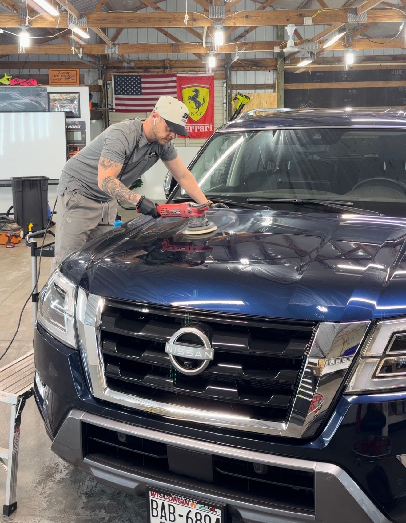A person in a gray shirt polishes the hood of a blue Nissan SUV inside a workshop with garage flags in the background.
