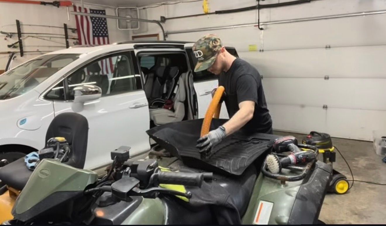 A person in a garage wears gloves while cleaning a black, rectangular item placed on top of a green all-terrain vehicle.