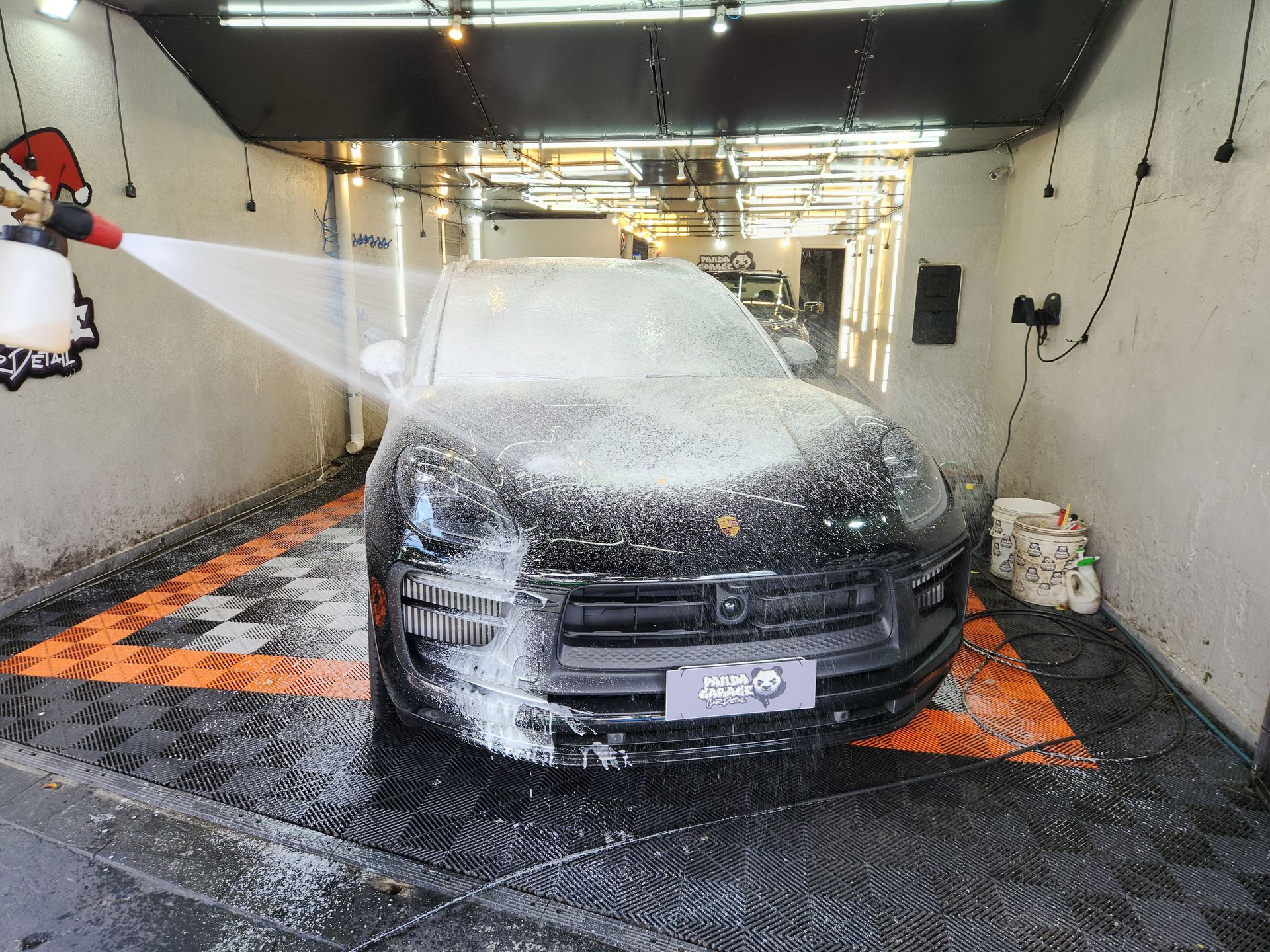 A black Porsche is being covered in white soap suds from a spray nozzle inside a brightly lit car wash bay.
