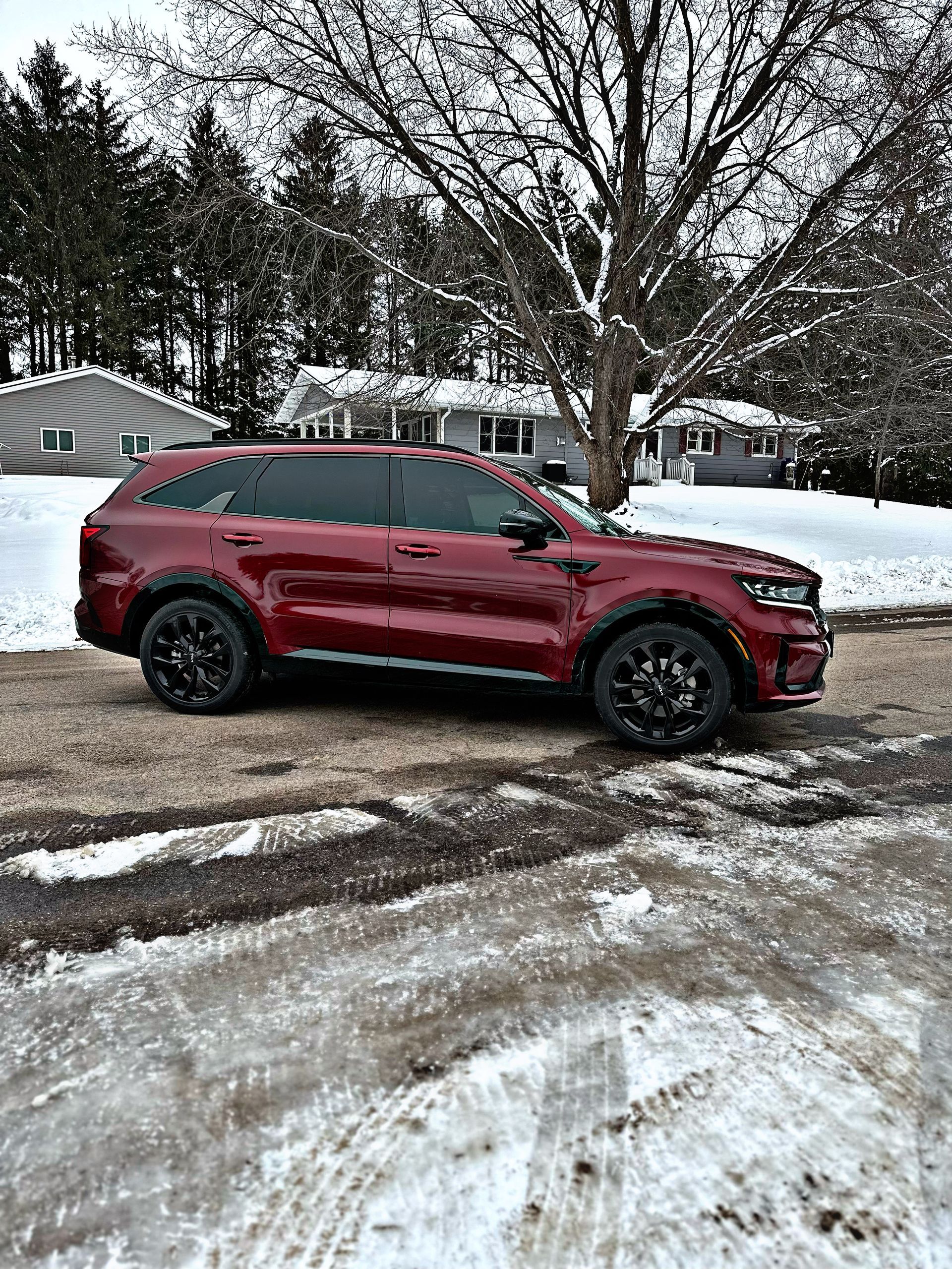 A dark red SUV parked on a gravel driveway covered in patches of snow with a house and trees in the background.