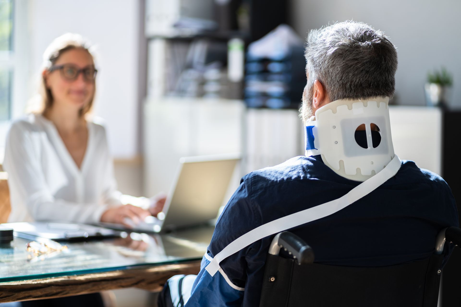 man wearing a neck brace and sitting in a wheelchair facing a woman at a desk with a laptop