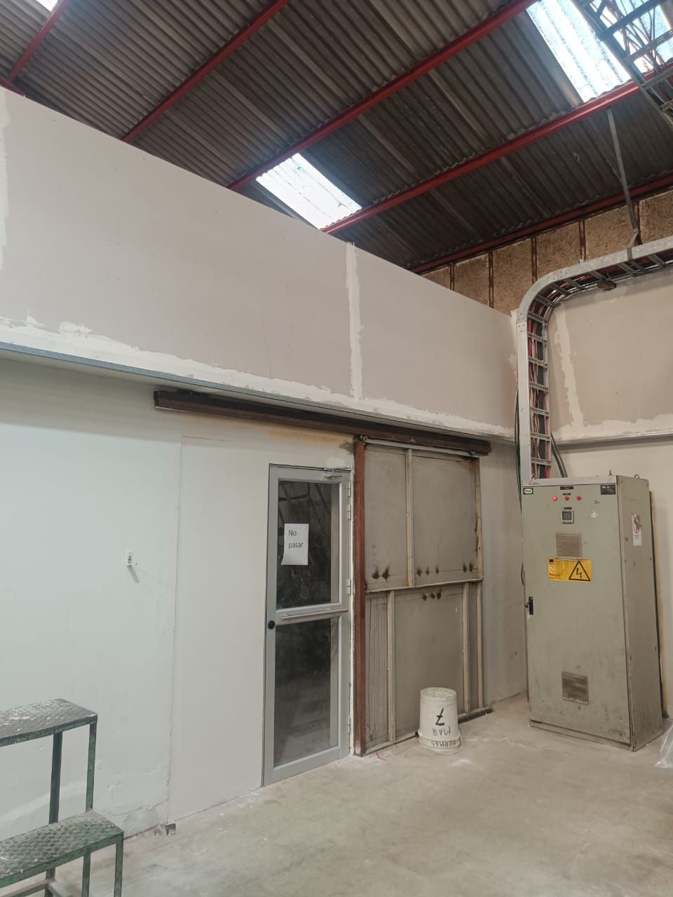 Habitación industrial con paredes blancas, puerta metálica, tuberías a la vista y tragaluces en un interior que recuerda a un almacén.
