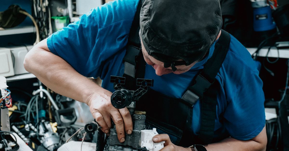 Mecánico con camisa azul inspeccionando un motor en un taller abarrotado de herramientas y maquinaria a su alrededor.