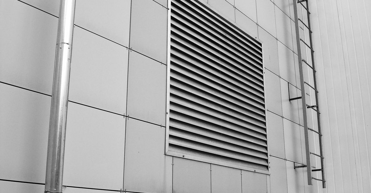 Pared de un edificio de metal gris con una gran rejilla de ventilación rectangular y tuberías verticales.