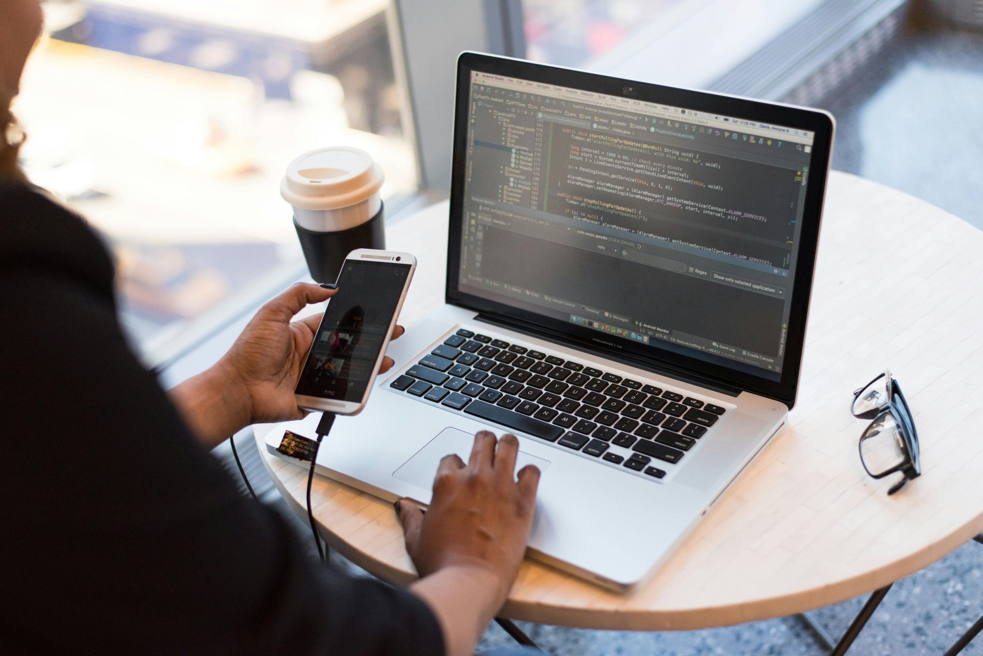 Person working on laptop, holding phone, coffee cup and glasses on table near window.