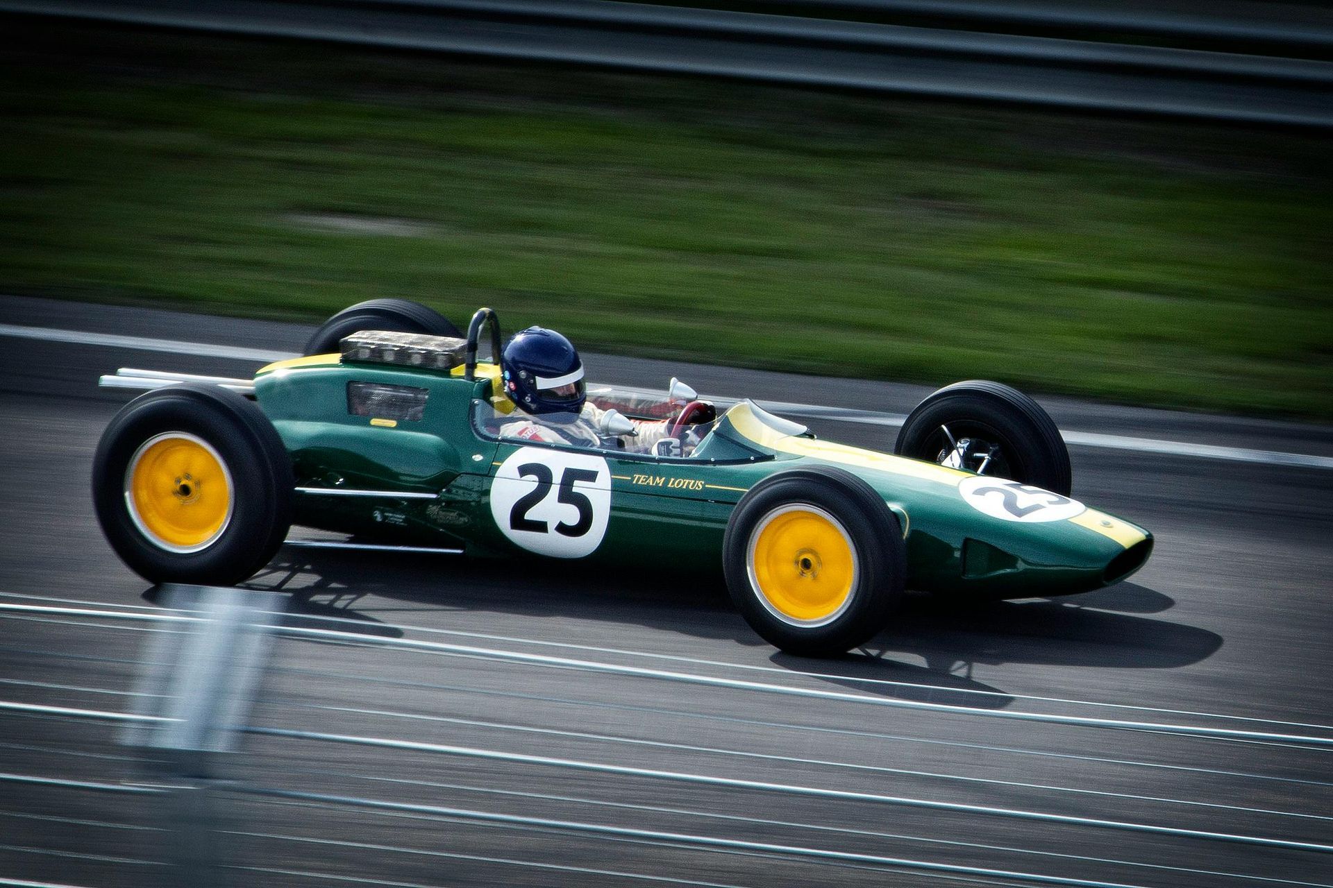 Green vintage race car, number 25, speeding on a track, driver in helmet.