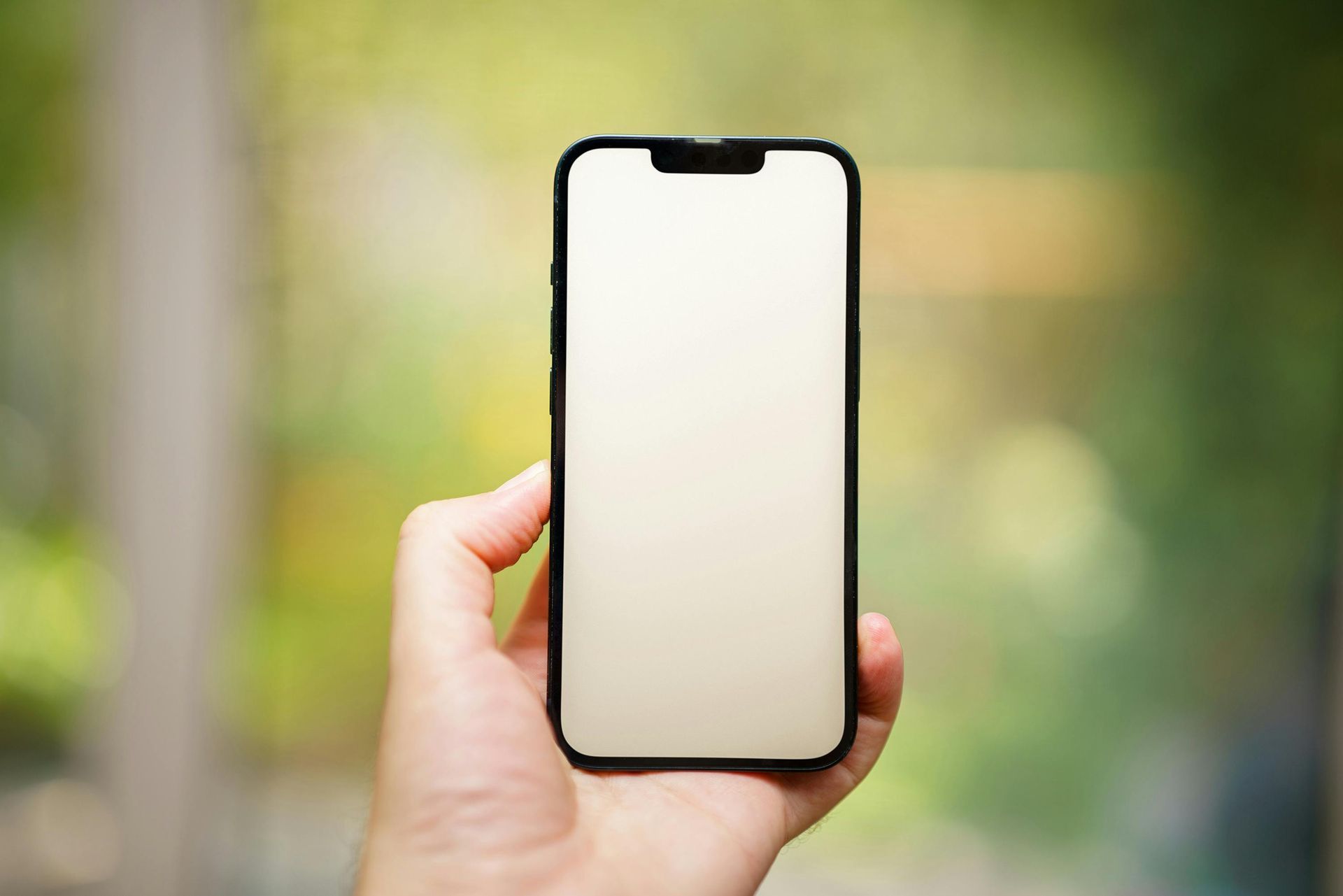 Hand holding a smartphone with a blank, light-colored screen against a blurry green background.