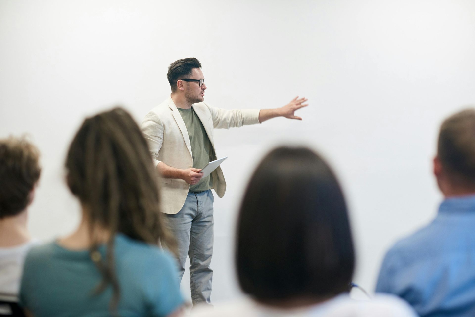 Man in light jacket gesturing during a presentation; audience seated in front of him.