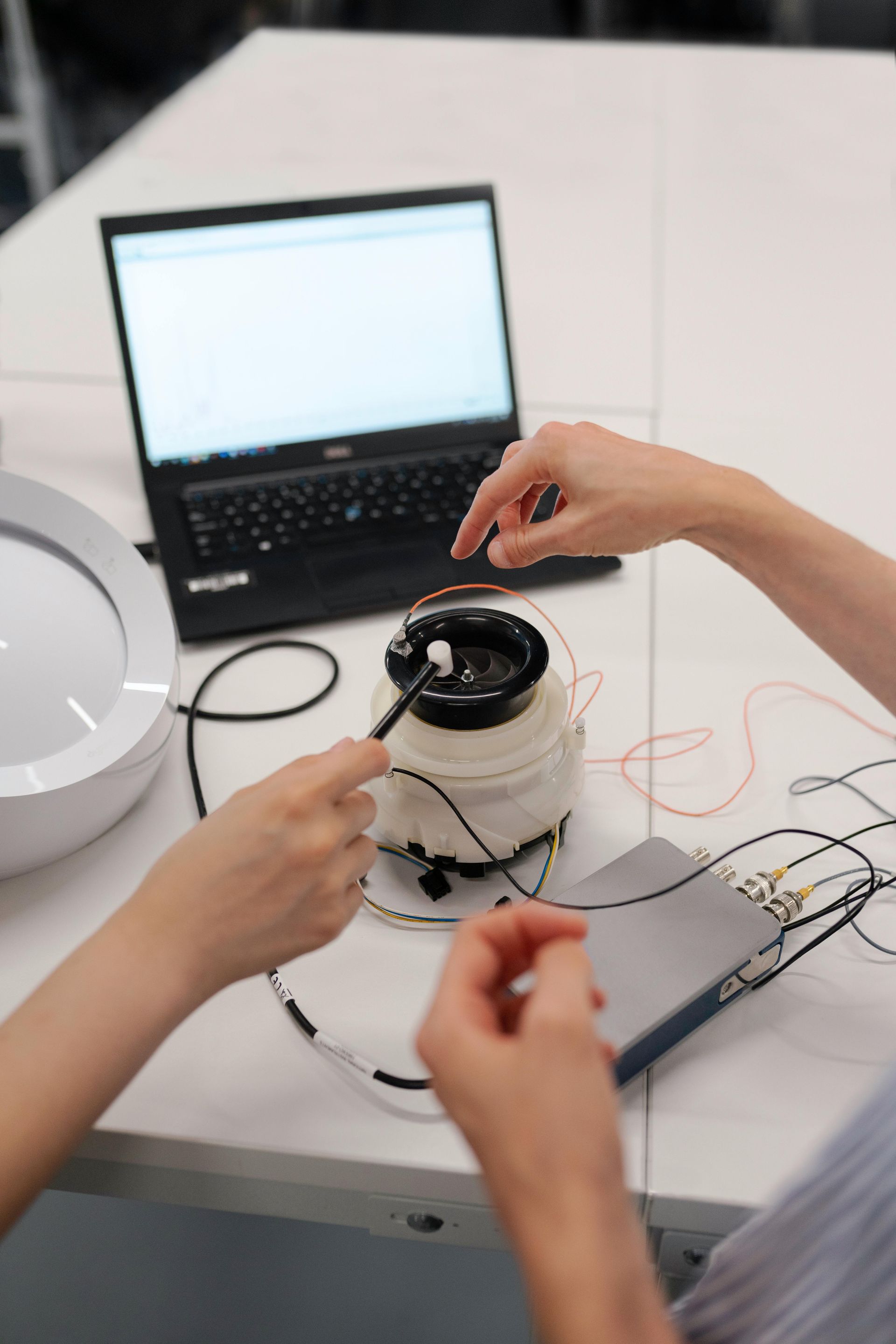 Two people working on a circuit. Wires connect to a jar-like object near a laptop and a white dish.