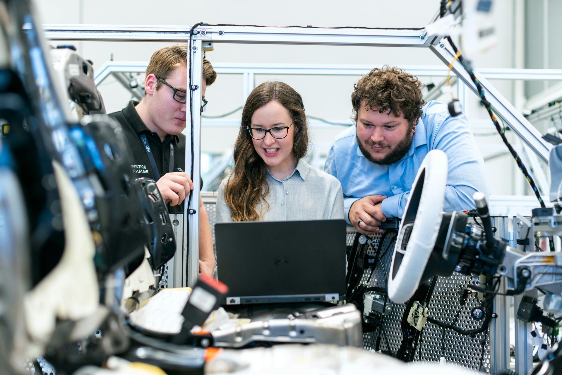 Engineers examine laptop in an automotive assembly setting; woman with glasses smiles, others look on.