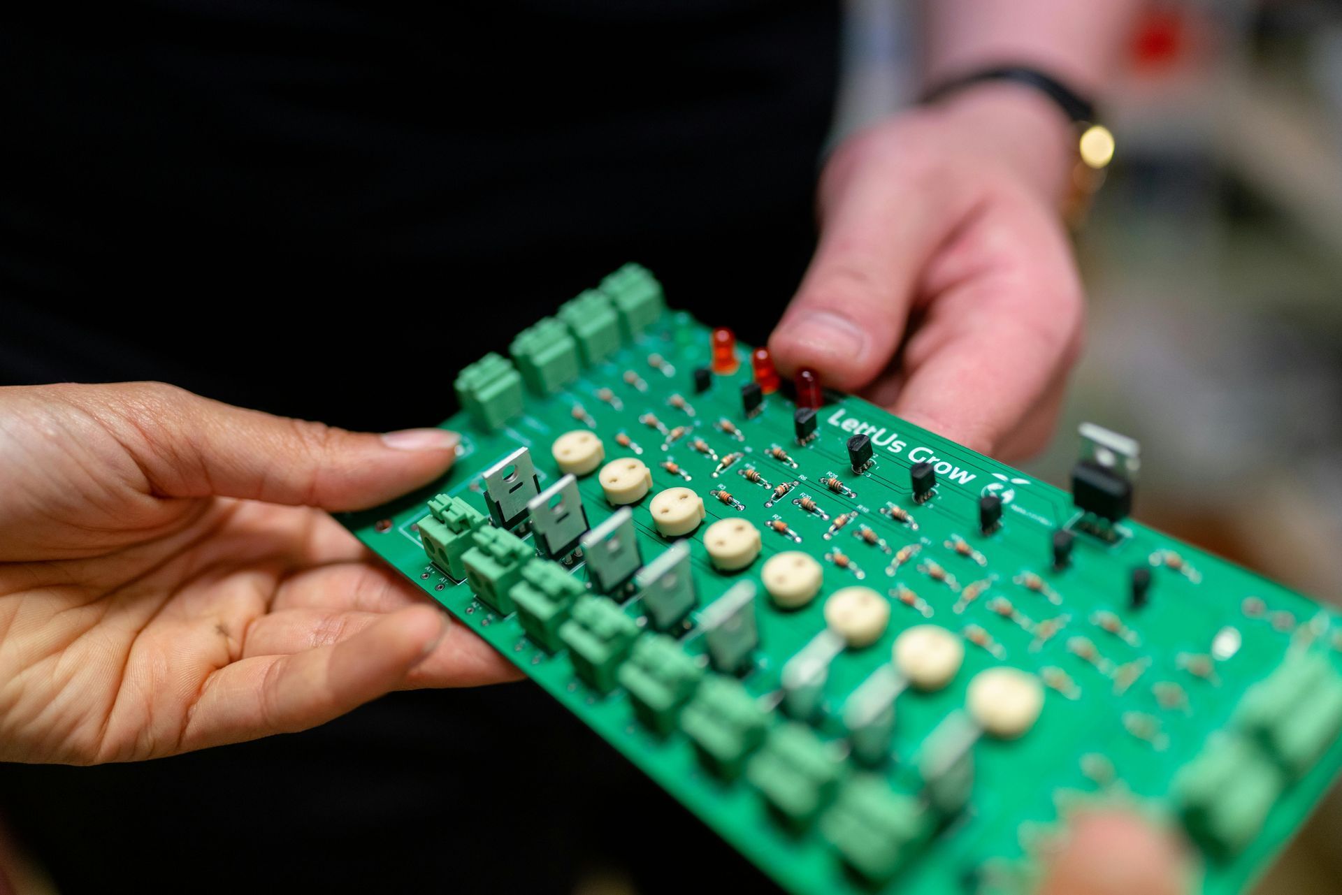 Hands holding a green circuit board with various components, including green connectors and red LEDs.