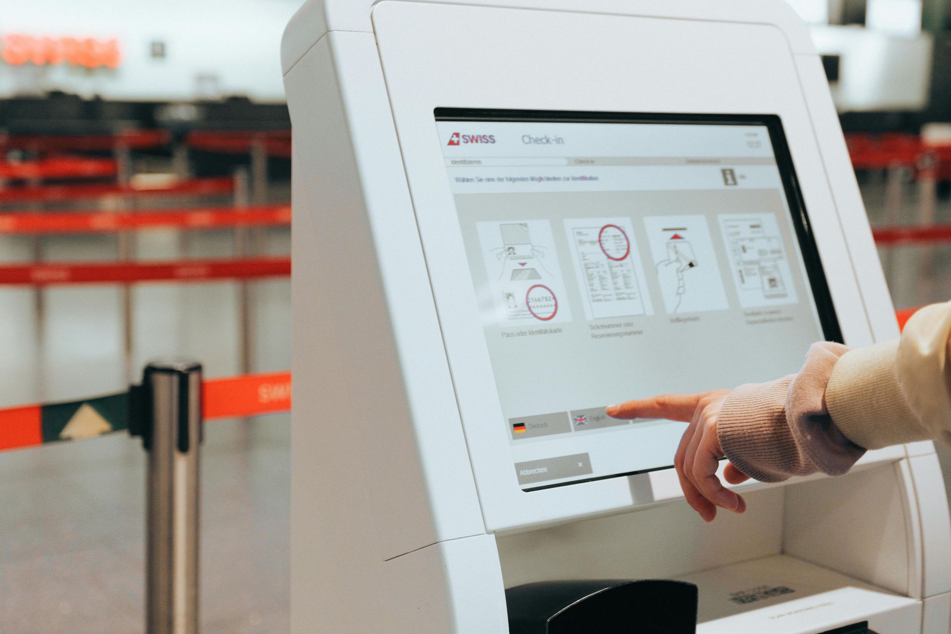 Person using a touchscreen airport check-in kiosk; white machine, red barriers in background.