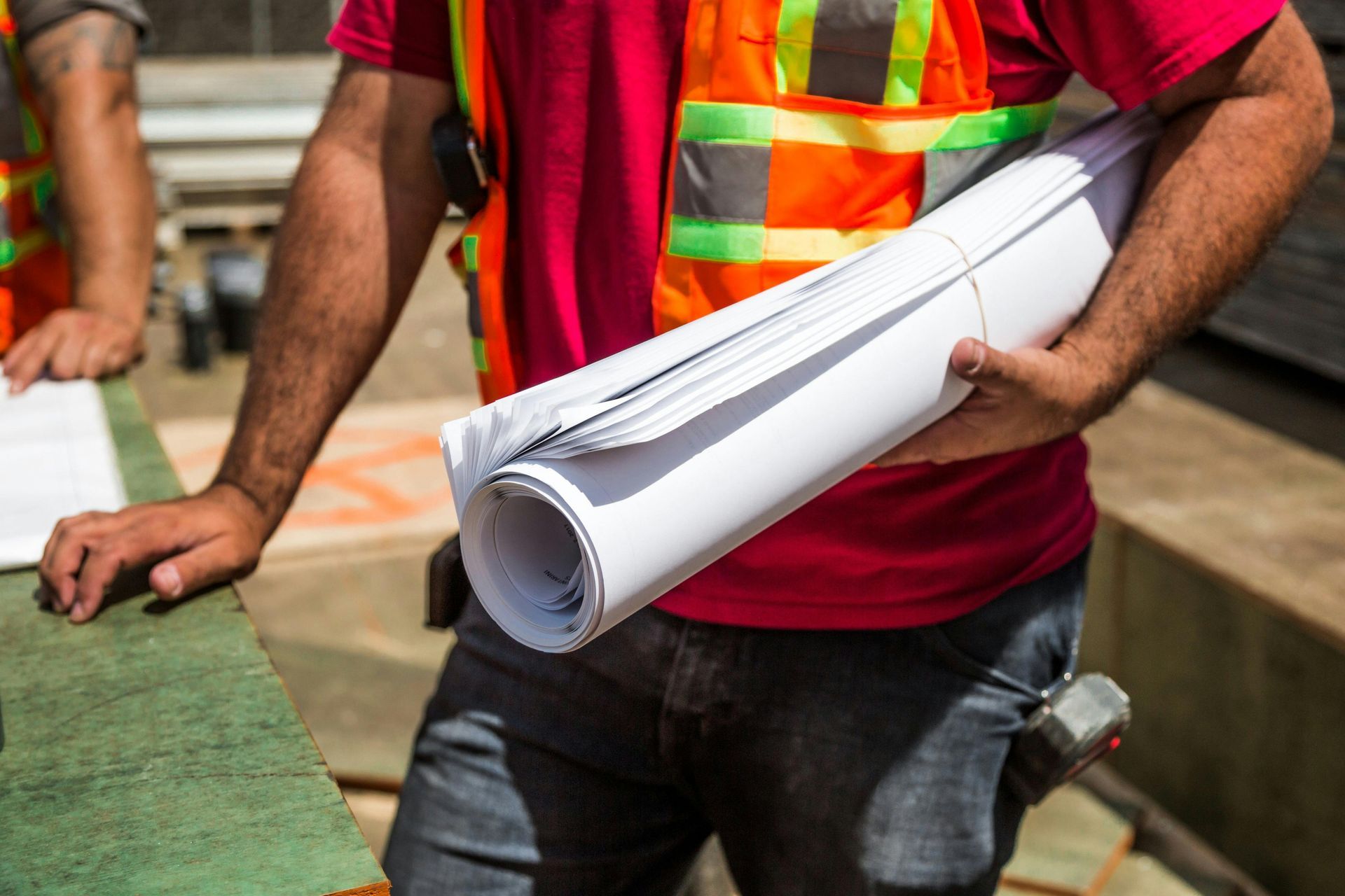 Construction worker in safety vest holding rolled blueprints at a construction site.
