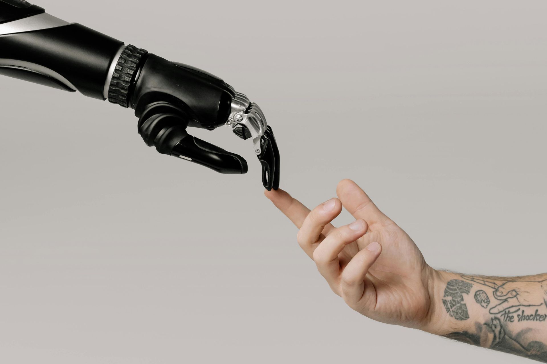Robot hand touches a human finger. Black robot arm, tattooed arm, gray background.