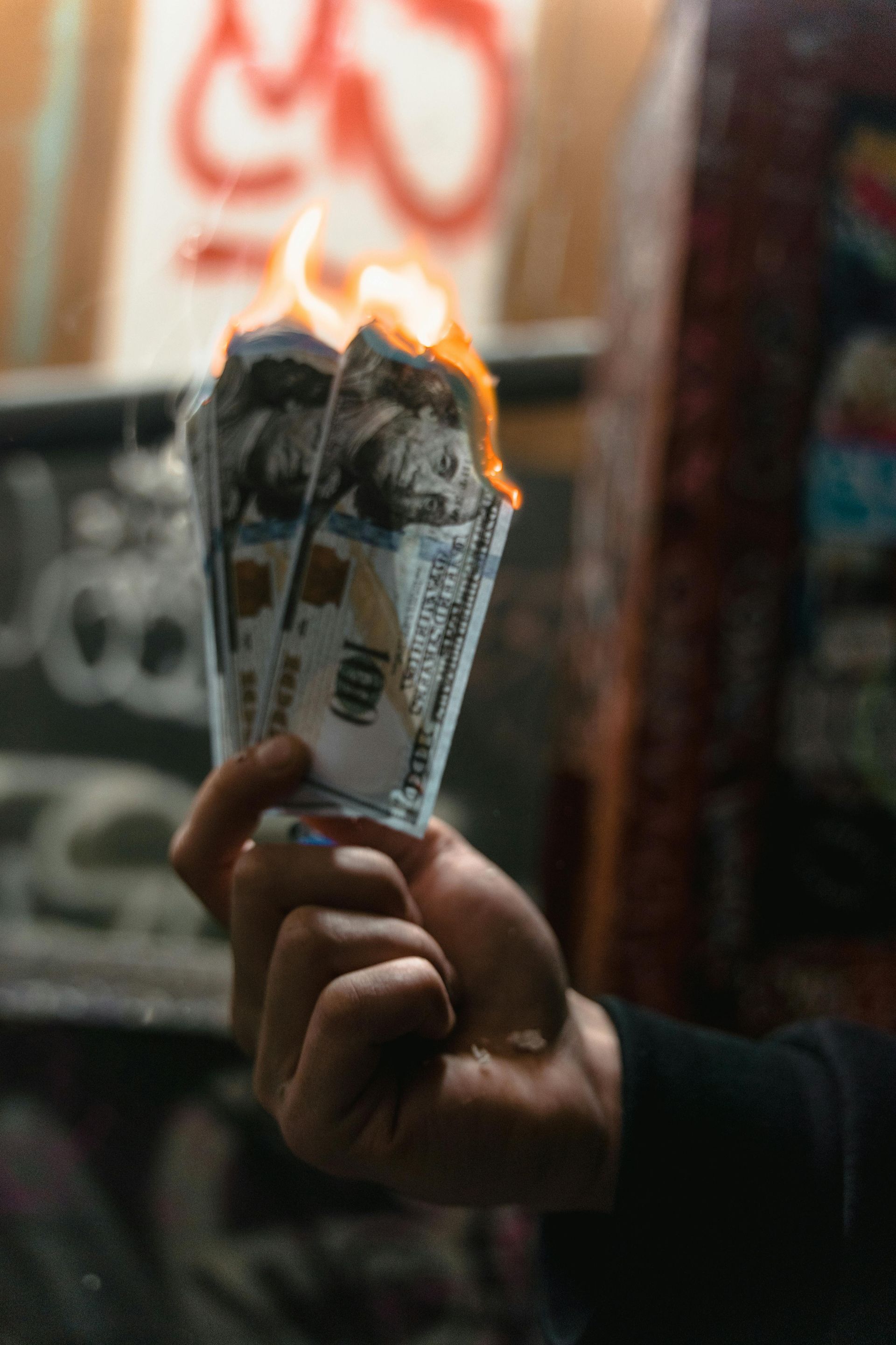 Hand holding burning $100 bills in front of a graffiti-covered wall. Flames visible.