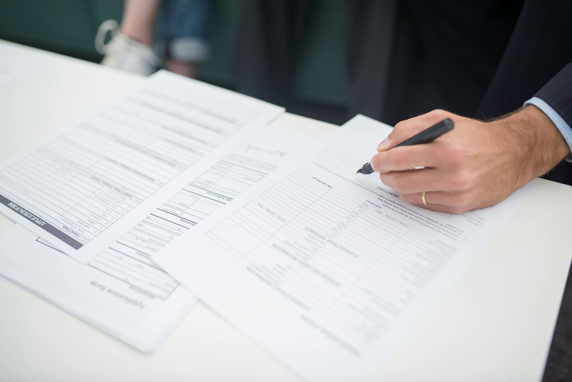 Person in suit signing paperwork with a pen on a white table.