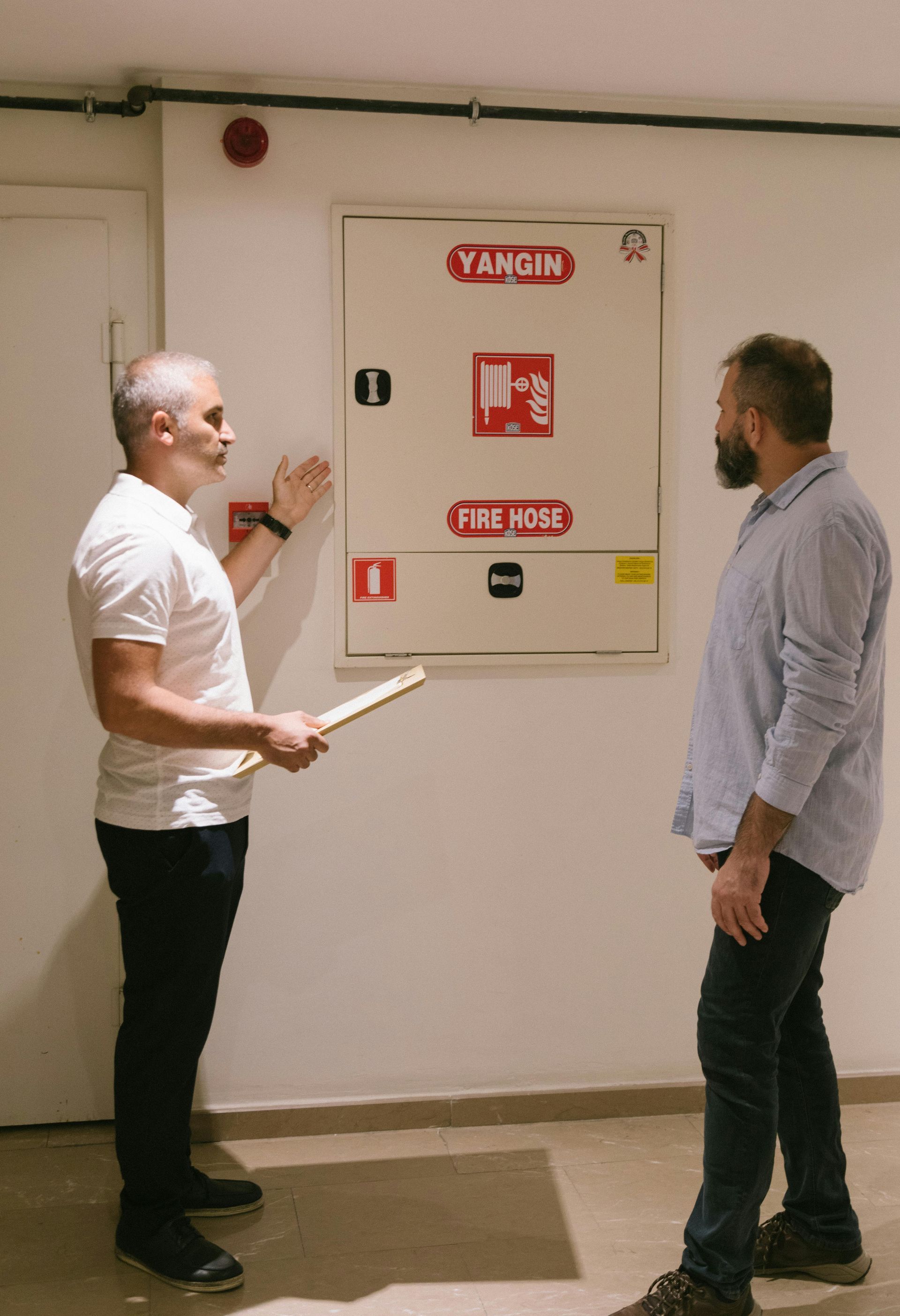 Two men by a fire hose cabinet; one points to it, other looks. Indoors, white walls, red signage.