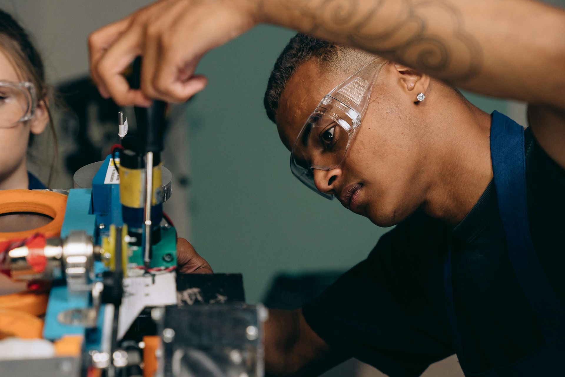 Man in safety glasses, using a screwdriver on machinery, focused expression. Workshop setting.