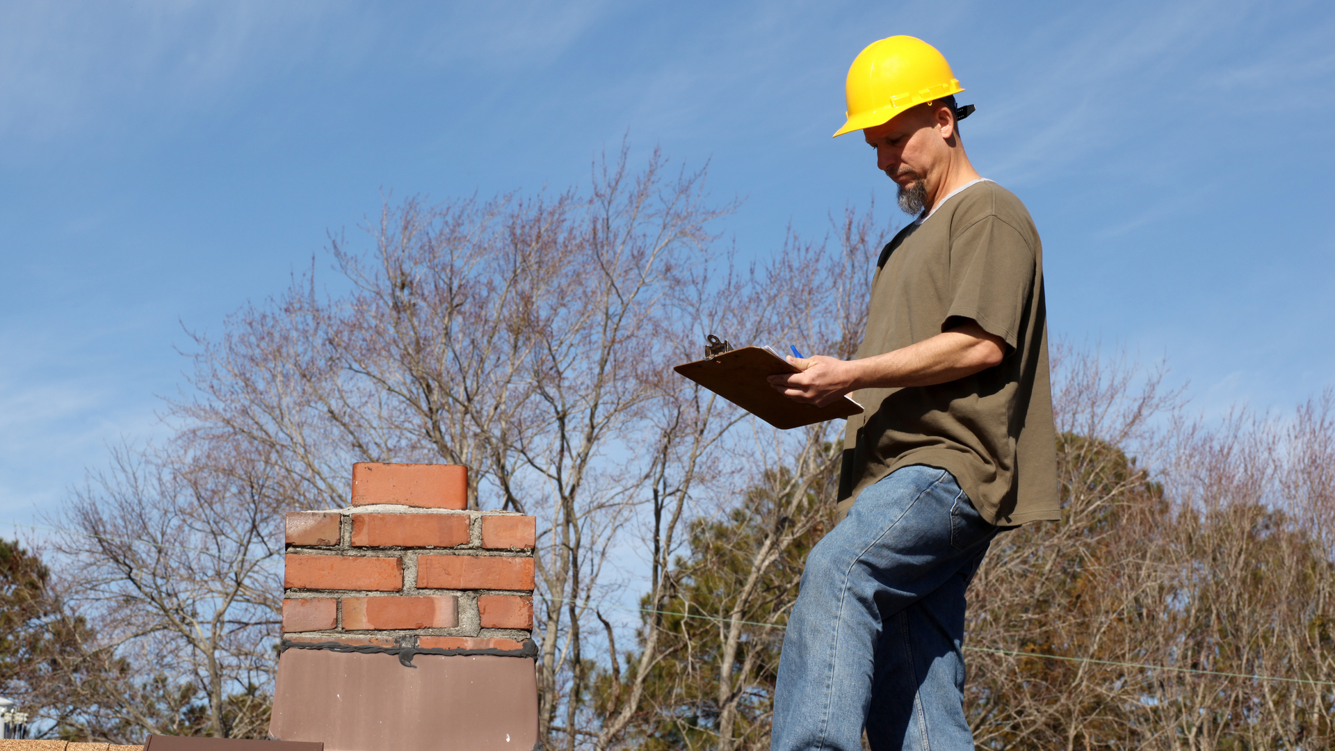 A man wearing a hard hat is standing on a roof looking at a clipboard.
