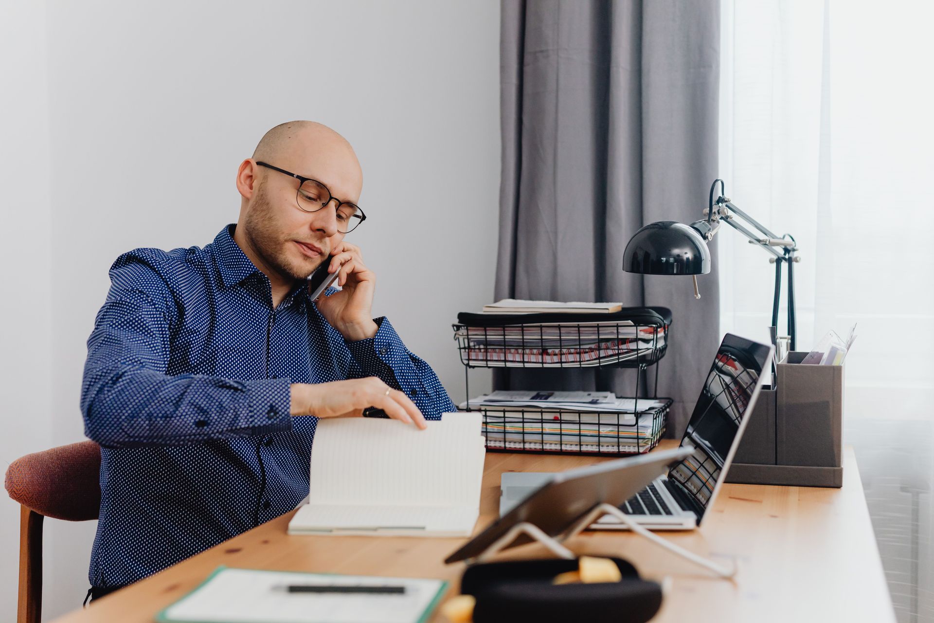 Man on phone at desk with laptop, papers, and desk lamp in a bright office