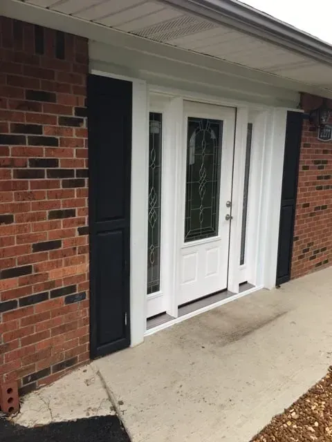 Brick house exterior with a white door, side windows, black shutters, and a concrete walkway.