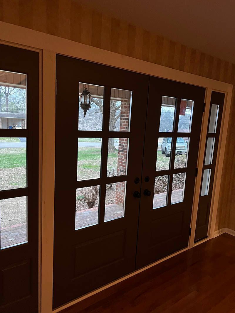 Brown French doors with glass panes and sidelight, framed in white, view to outside.