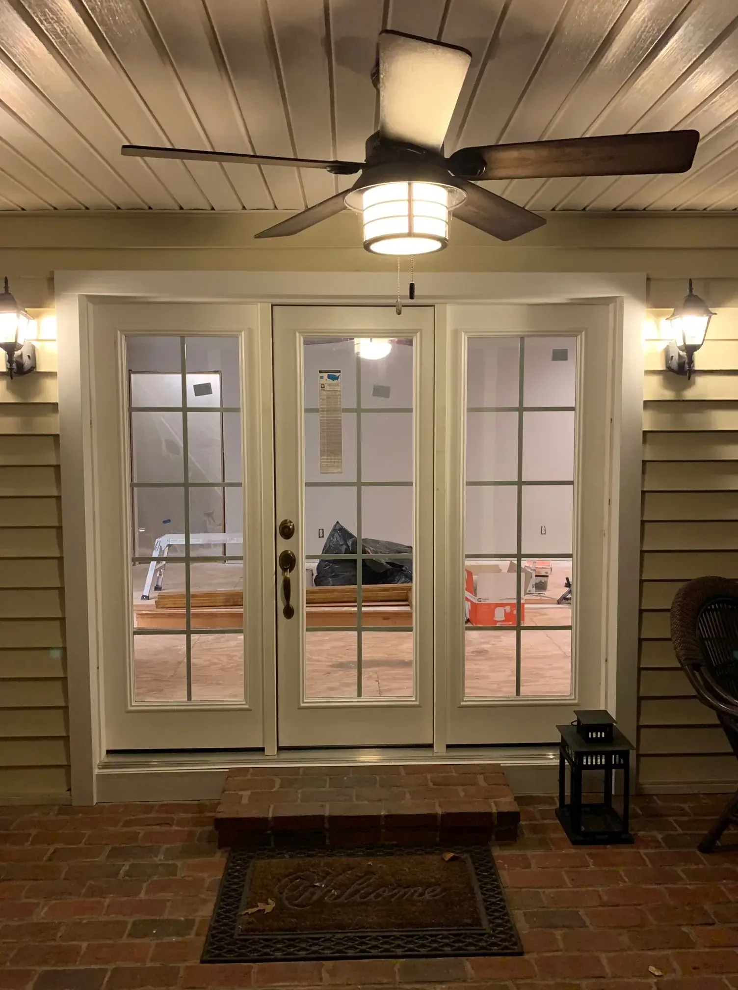Three white-framed French doors, with brick step, opening to interior room under construction. Overhead fan and wall-mounted lights.