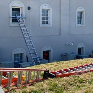 A tall ladder leans against a building's window. Another ladder rests in the grass. Air conditioning units sit below windows.