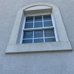 Window with white frame and blinds, set into a stucco wall with a curved top and concrete trim.