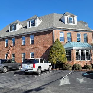 Brick building with multiple windows, a grey roof, and parked cars on a sunny day.