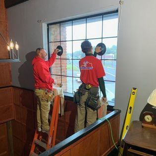Two workers installing a window. One on a ladder, using suction cups. The other is using suction cups as well.