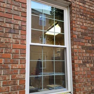 White-framed window in a red brick wall, reflecting an interior room with a desk and overhead light.