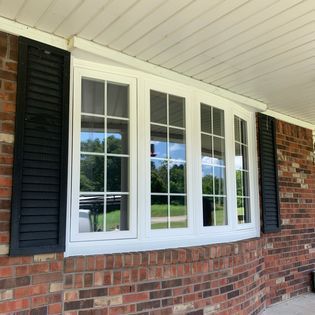 White-framed, multi-pane bay window with black shutters on a brick building; reflections of trees and sky visible.