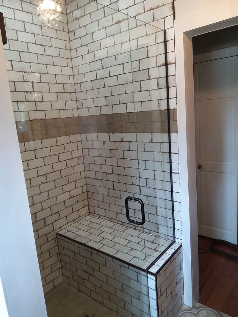 Tiled shower with a built-in bench. The walls are white brick-pattern tiles, with a horizontal brown stripe. A glass door is present.