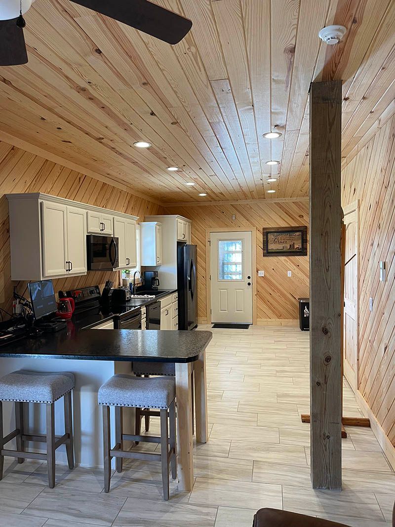 Kitchen with wood-paneled walls and ceiling, white cabinets, black countertops, and a stone floor; two stools at the island.