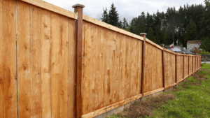 Wooden fence, stained light brown, runs along a grassy area towards distant trees and a cloudy sky.