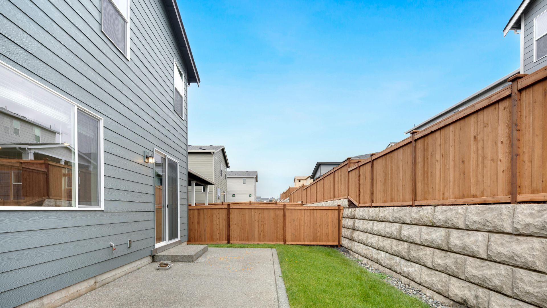 Backyard view of a house with gray siding, a concrete patio, and a wooden fence along a retaining wall.