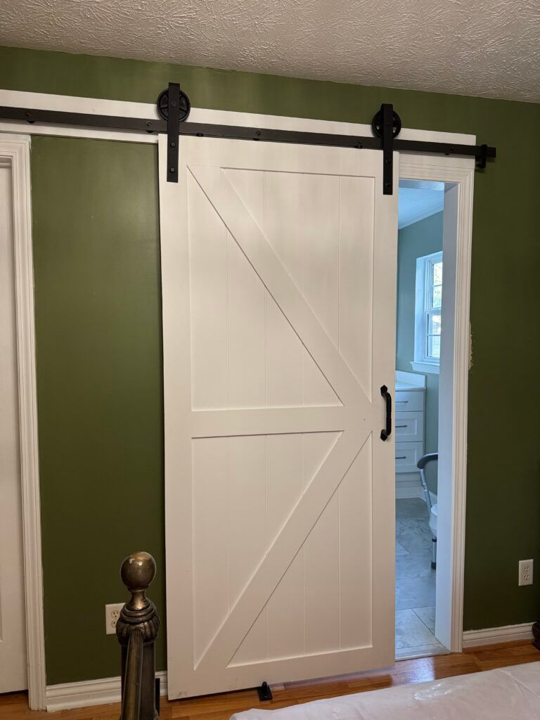 White barn door with black hardware, hanging on a track in a room with green walls.